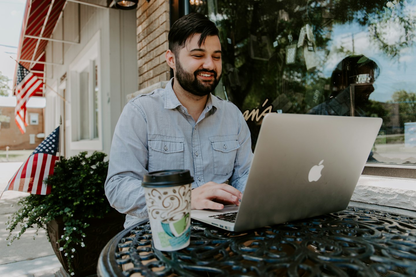 Man smiles working on laptop at outdoor cafe with coffee.